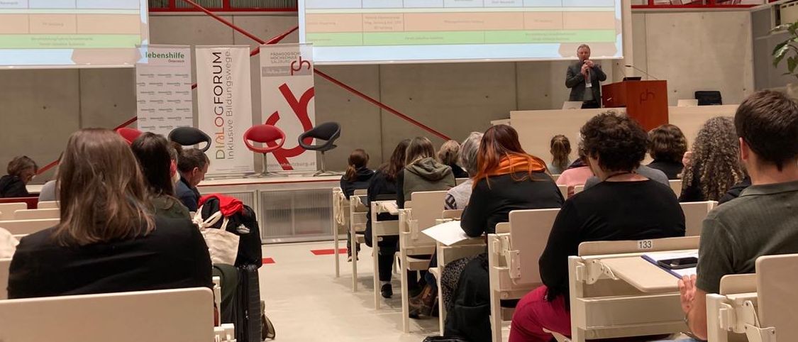 Ein Mann spricht an einem Podium mit einer Projektionsleinwand hinter ihm. Ein Publikum sitzt in Stühlen vor ihm.