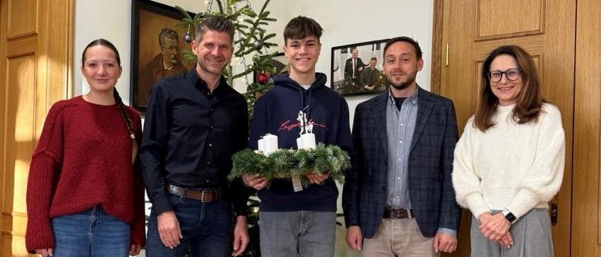 Fünf Personen stehen eng beieinander in einem Raum und lächeln. Der Mann in der Mitte hält eine Girlande. Dahinter steht ein Weihnachtsbaum und es hängen Bilderrahmen an der Wand.