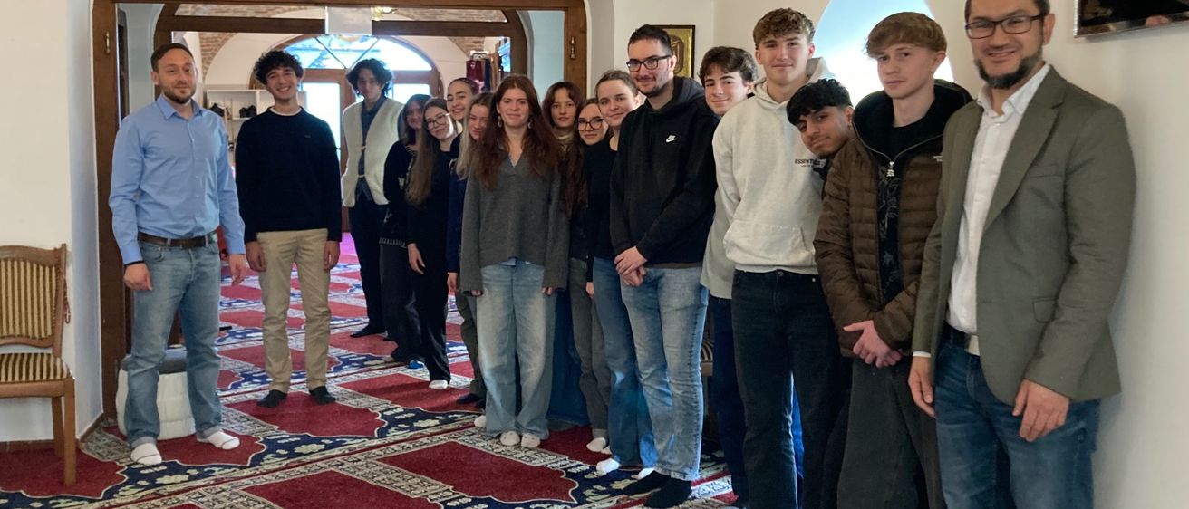 Eine Gruppe von Menschen steht in einem Raum, vielleicht einer Moschee, mit Teppichen auf dem Boden. Eine Kronleuchter hängt von der Decke.