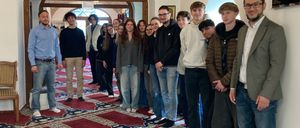 Eine Gruppe von Menschen steht in einem Raum, vielleicht einer Moschee, mit Teppichen auf dem Boden. Eine Kronleuchter hängt von der Decke.
