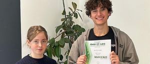 Zwei junge Leute stehen vor einer Wand und halten Urkunden in den Händen. Das Mädchen trägt einen schwarzen Rock und der Junge eine graue Jacke. Sie lächeln beide. Hinter ihnen steht ein lila Sofa mit einem weißen Stuhl und einer großen Pflanze.