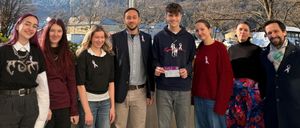 Eine Gruppe von Menschen posiert für ein Foto vor einer rosa Flagge vor einer Bergkulisse. Sie tragen rosa Schleifen und haben lächelnde Gesichter.