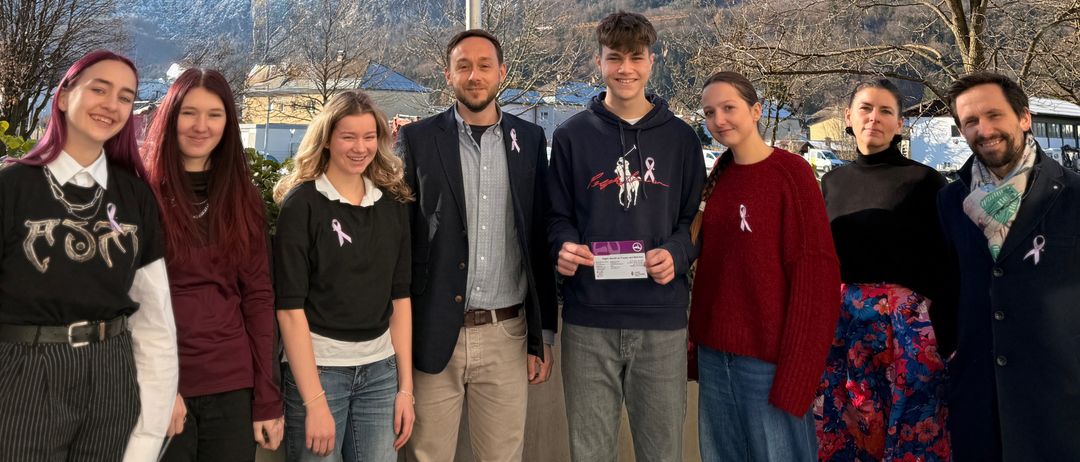 Eine Gruppe von Menschen posiert für ein Foto vor einer rosa Flagge vor einer Bergkulisse. Sie tragen rosa Schleifen und haben lächelnde Gesichter.