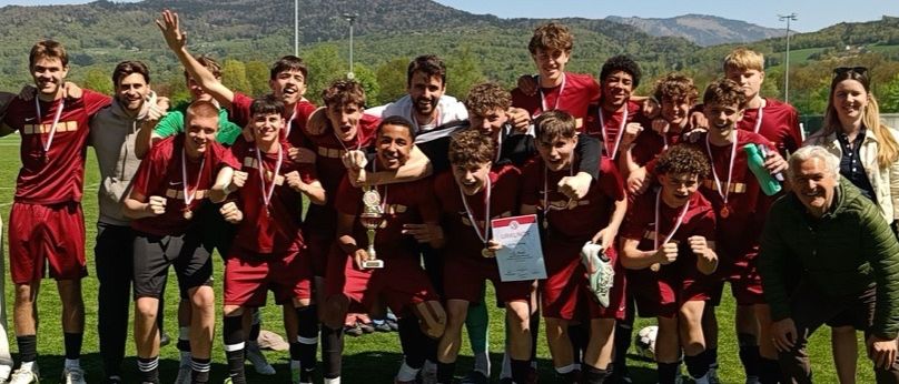Eine Fußballmannschaft steht auf einem Feld, feiert mit Medaillen um den Hals, hält einen Pokal und trägt rote Uniformen. Im Hintergrund gibt es Berge und einen klaren Himmel.