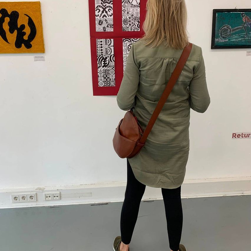 Eine Frau steht vor einer Wand mit mehreren Kunstwerken. Sie hat eine braune Schultertasche. An der Wand befindet sich eine Steckdose und ein Schild mit der Aufschrift 'Retur'.