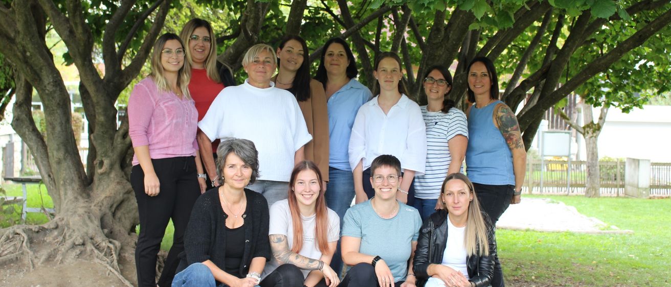 Eine Gruppe von Frauen in verschiedenen Outfits, die unter einem großen Baum mit grünen Blättern posiert, einige mit Brille und lächelnd.
