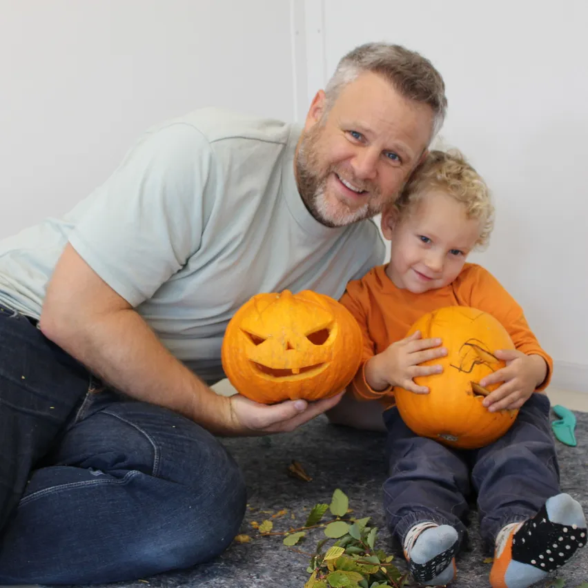 Ein Vater und sein Sohn lächeln und halten Kürbisse. Der Vater hat einen geschnitzten Kürbis und der Sohn einen Kürbis mit einem Gesicht. Sie sitzen auf dem Boden.