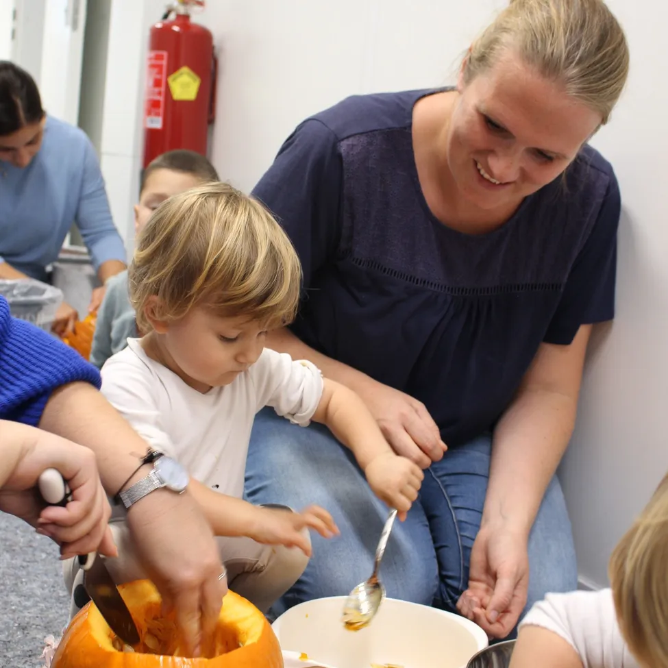 Eine Frau hilft einem Kind, einen Kürbis mit einem Messer zu schnitzen, während ein weiteres Kind und ein Erwachsener zusehen. Ein Feuerlöscher ist an der Wand hinter ihnen montiert.