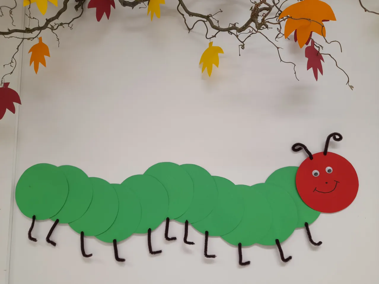 Eine handgefertigte Papierraupe mit grünen Körpersegmenten und einem roten Kopf, die von einem Baumzweig mit Herbstblättern hängt.