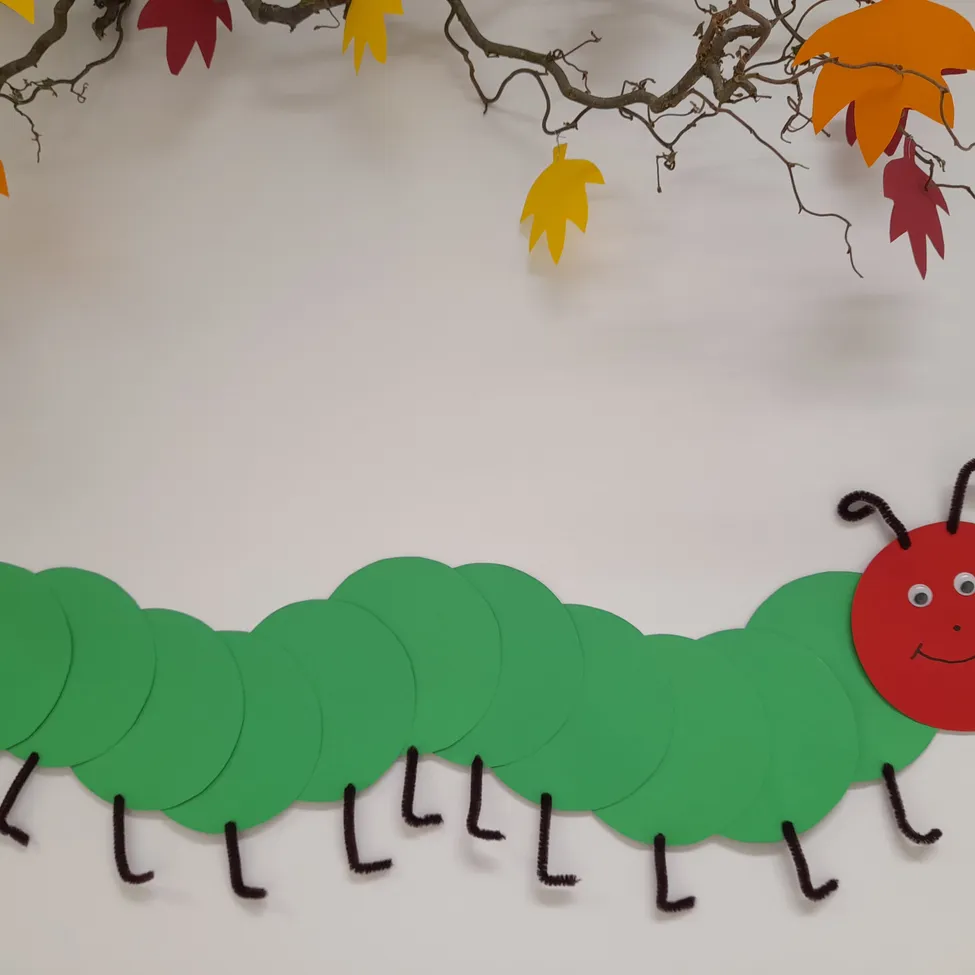 Eine handgefertigte Papierraupe mit grünen Körpersegmenten und einem roten Kopf, die von einem Baumzweig mit Herbstblättern hängt.