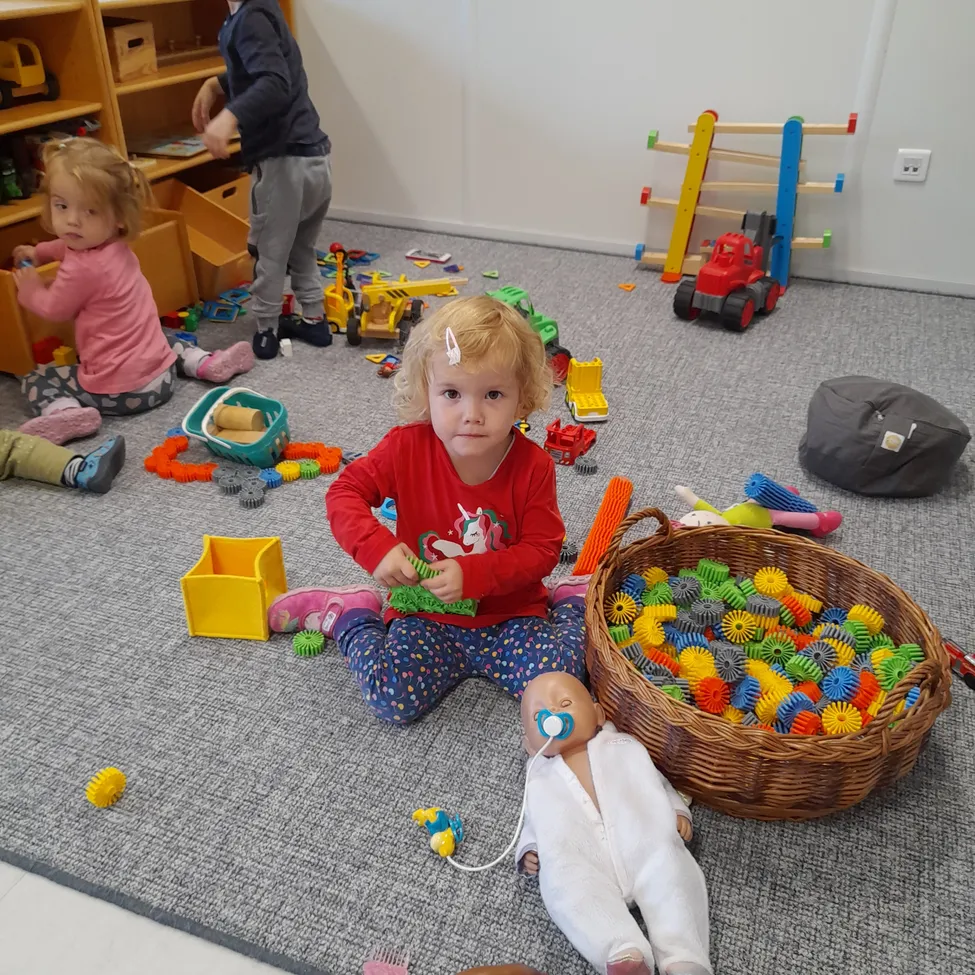 Ein kleines Mädchen sitzt auf dem Boden, umgeben von Spielzeug, darunter ein Korb mit bunten Zahnrädern. Ein anderes Kind spielt in der Nähe.