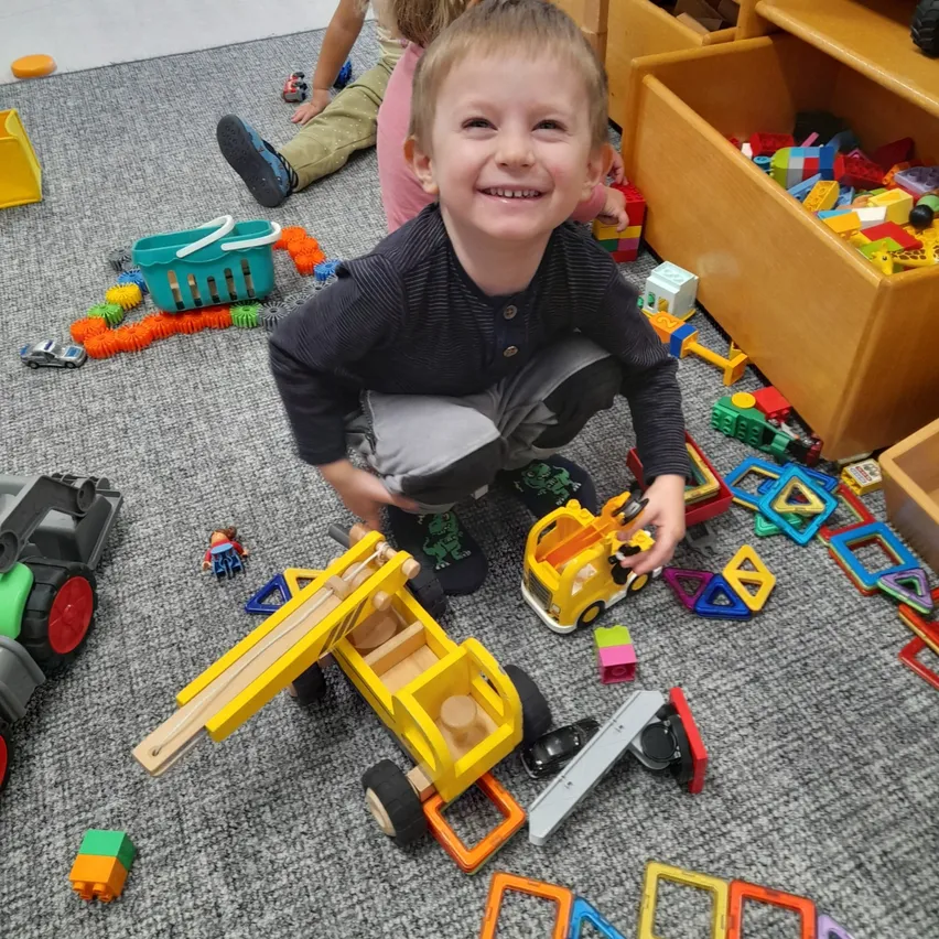 Ein kleiner Junge spielt mit Spielzeugautos und Bausteinen auf einem Teppichboden. Er lächelt und hält einen gelben Spielzeuglastwagen.