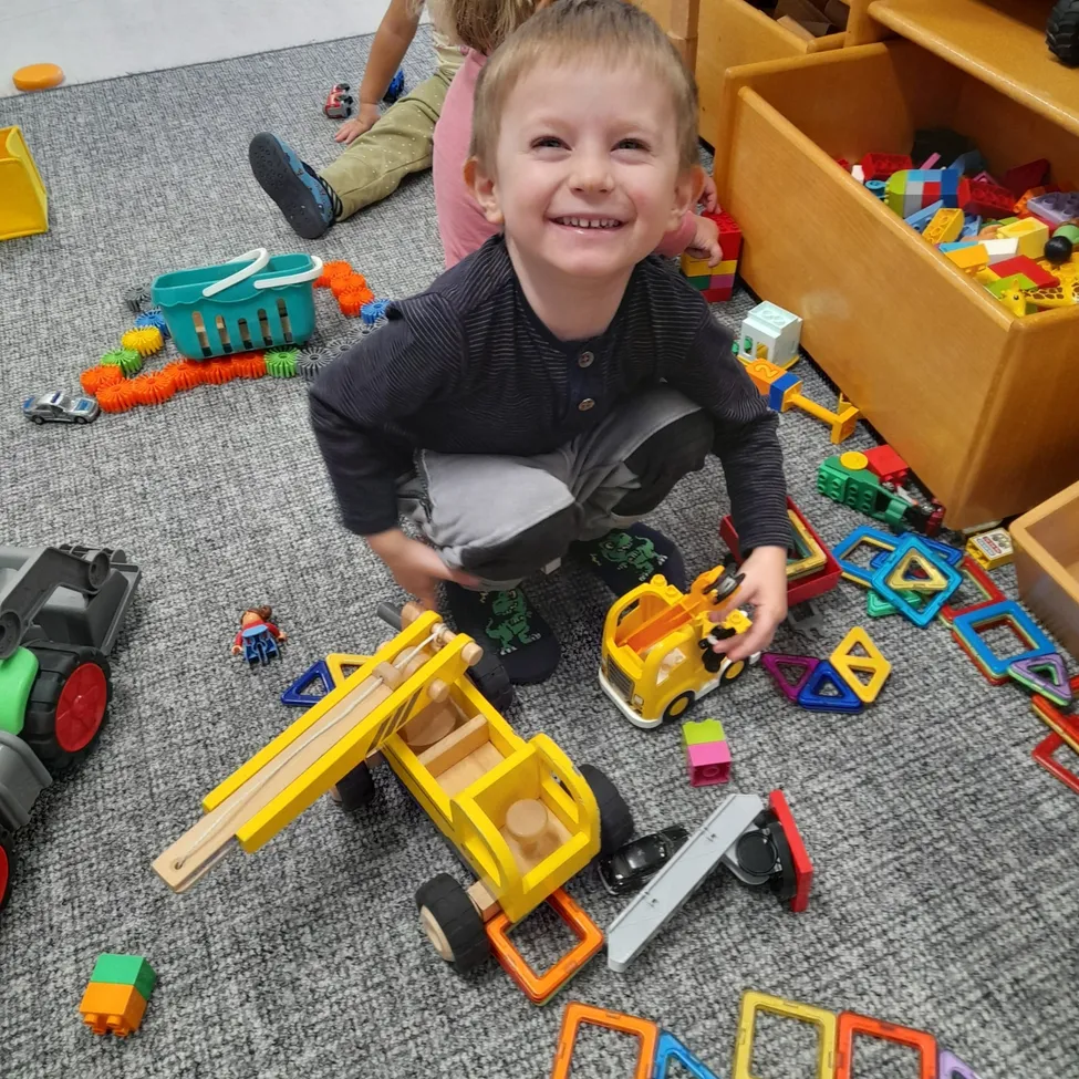 Ein kleiner Junge spielt mit Spielzeugautos und Bausteinen auf einem Teppichboden. Er lächelt und hält einen gelben Spielzeuglastwagen.