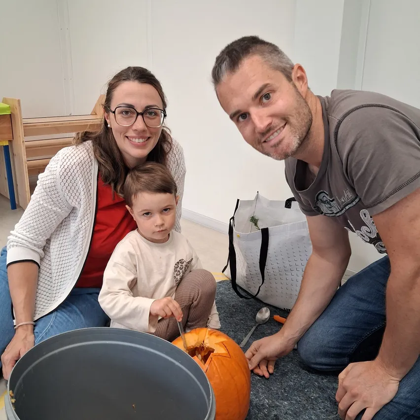 Eine Familie sitzt auf dem Boden, lächelt und schnitzert eine Kürbis. Die Frau trägt eine Brille und einen Armreif, und der Mann hält einen Löffel. Das Kind hält ein Messer und ist dabei, den Kürbis zu schneiden. Es gibt einen Eimer und eine Tasche auf dem Boden.