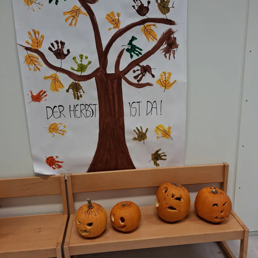 Ein Poster mit einem Baum und Handabdrücken hängt an einer Wand. Darunter steht eine Holzbank mit vier geschnitzten Kürbissen.