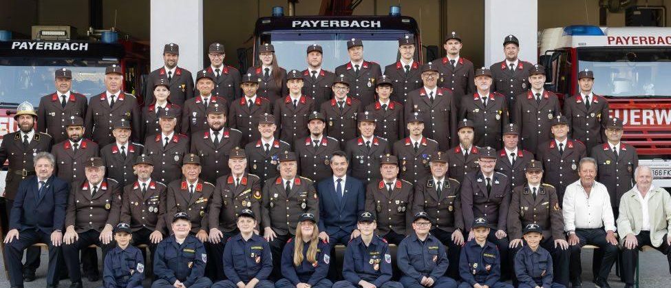 Das Feuerwehrteam von Payerbach posiert für ein Gruppenfoto vor ihrer Feuerwache. Sie tragen alle Uniformen, einige tragen Hüte. Hinter ihnen sind Feuerwehrfahrzeuge geparkt.