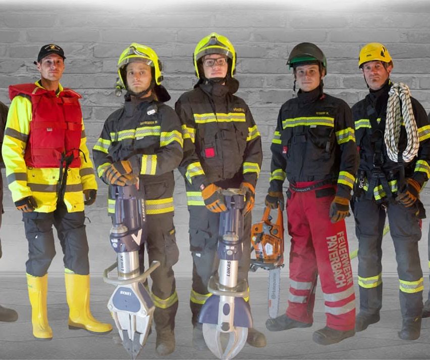Fünf Feuerwehrleute in Uniform stehen vor einer Ziegelwand, jeder hält verschiedene Werkzeuge. Einer hält eine Kettensäge, ein anderer hat ein großes Werkzeug, während andere kleinere Werkzeuge und Seile haben.