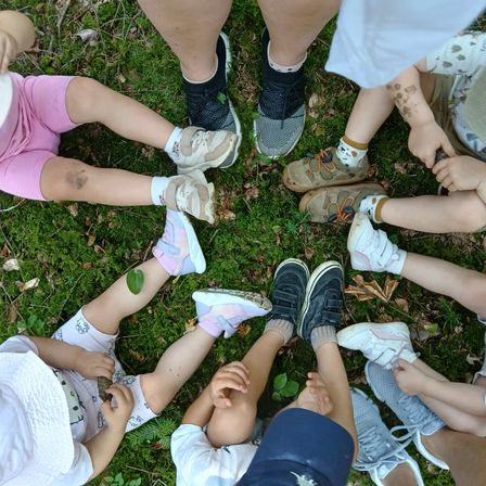 Eine Gruppe von Kindern und Erwachsenen sitzt im Kreis auf dem Gras, einige mit Schmutz auf ihren Kleidern und Schuhen.