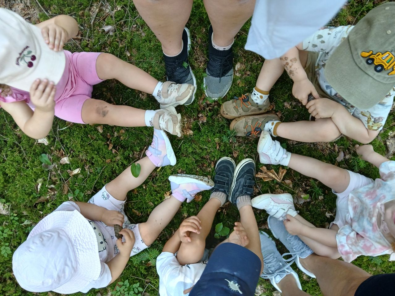 Eine Gruppe von Kindern und Erwachsenen sitzt im Kreis auf einem Grasfeld, einige mit Schmutz an ihren Kleidern und Schuhen.