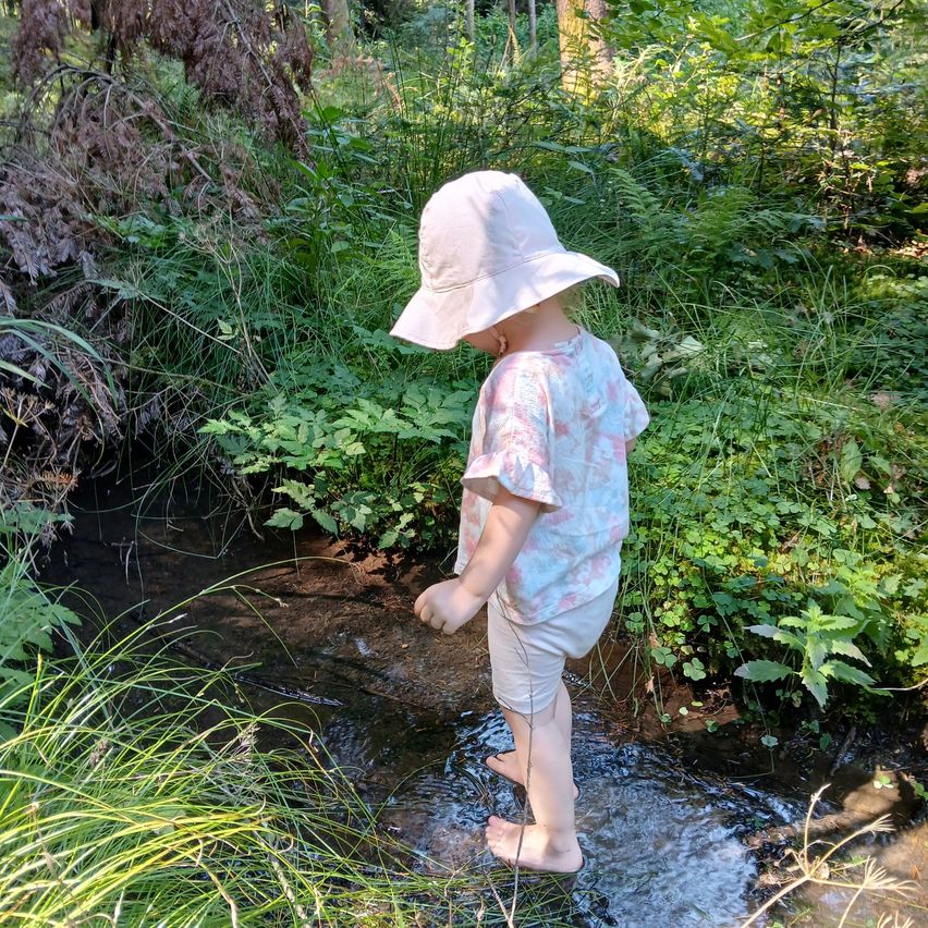 Ein kleines Kind mit Hut und Shorts steht in einem kleinen Bach, umgeben von üppiger Vegetation.