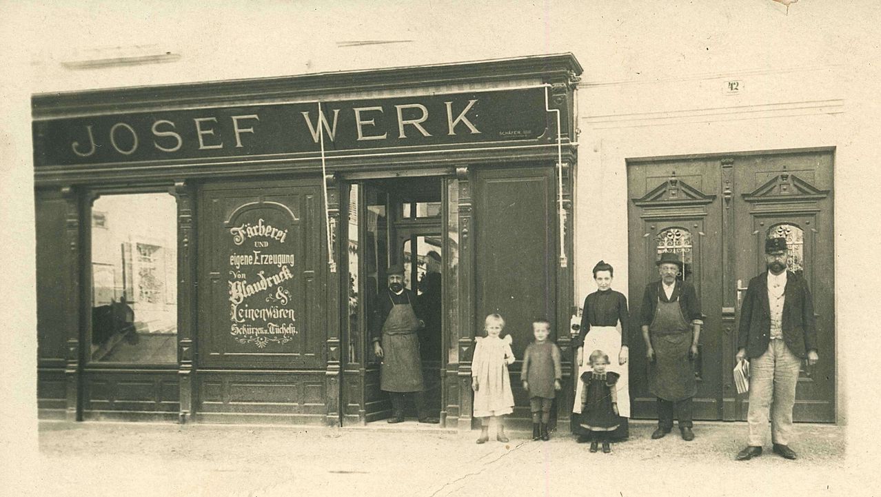 Ein altes Foto einer Familie vor einem Geschäft namens EF WERK, mit Schildern für Stoffe und Leinen.