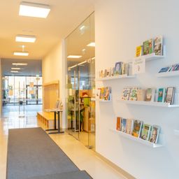 Ein Korridor mit weißen Wänden und Glaswänden und einem Teppich. Bücher sind auf weißen Regalen ausgestellt. Der Korridor führt zu einem Ausgang mit einer Glastür.