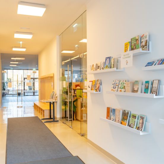 Ein Korridor mit weißen Wänden und Glaswänden und einem Teppich. Bücher sind auf weißen Regalen ausgestellt. Der Korridor führt zu einem Ausgang mit einer Glastür.