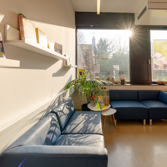 Ein helles Zimmer mit einer blauen Couch, einem kleinen Tisch und einem Fenster mit Blick auf Bäume und eine Kirche.