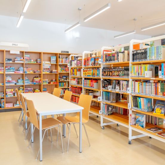 Eine leere Bibliothek mit mehreren Tischen, Stühlen und Bücherregalen, die mit Büchern und Brettspielen gefüllt sind. Jedes Regal hat beschriftete Abschnitte.