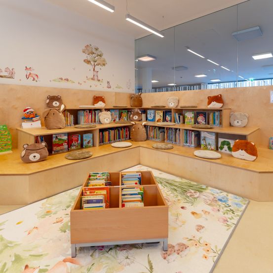 Eine Kinderbibliothek mit einer geschwungenen Sitzecke. Gefüllte Tiere und Bücher stehen auf den Regalen, mit einer kleinen Holzkiste auf dem Boden.