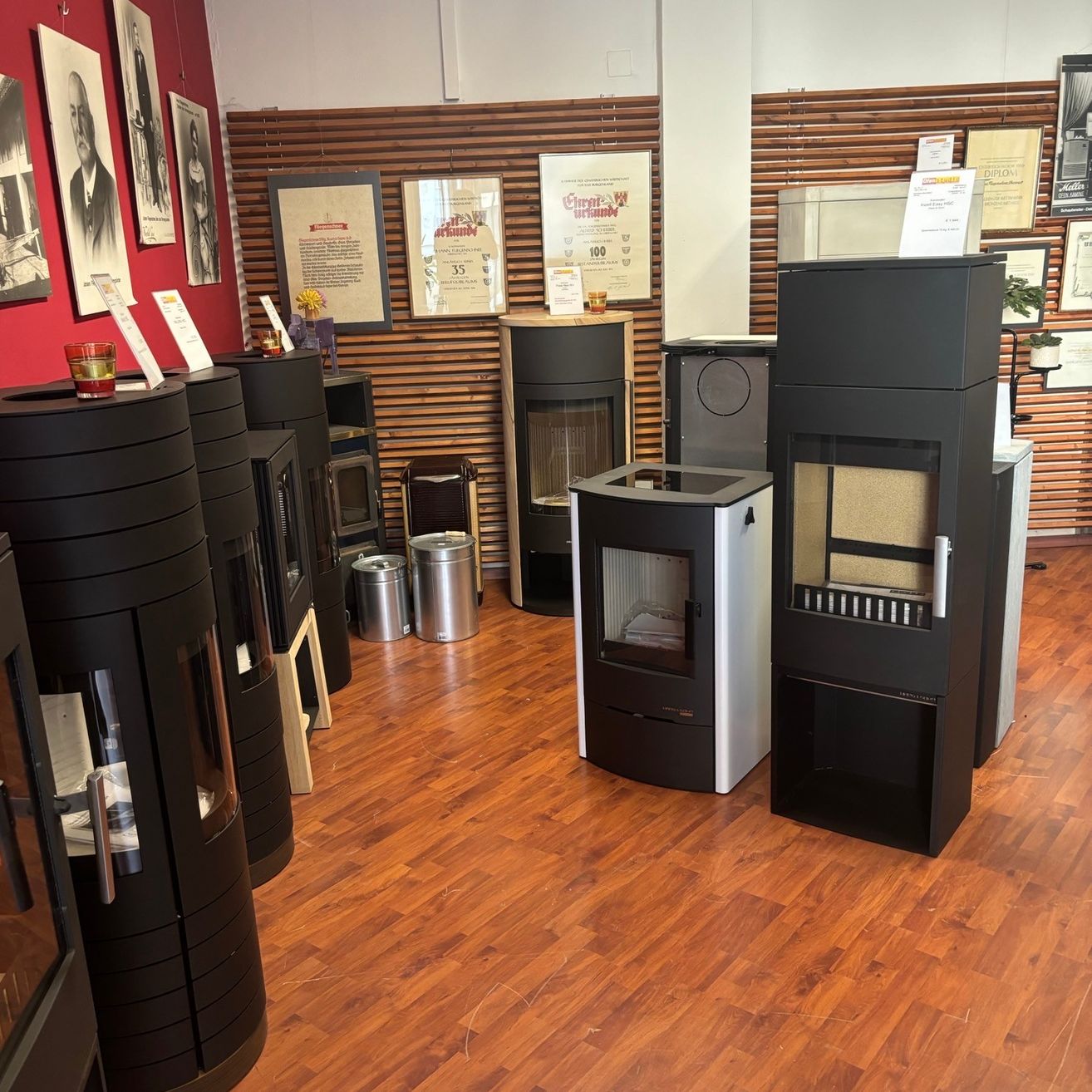 Ein Ausstellungsraum mit Holzboden zeigt verschiedene schwarze und weiße Kamine. Mehrere Mülleimer sind um den Raum herum aufgestellt, und mehrere gerahmte Bilder schmücken die Wände.