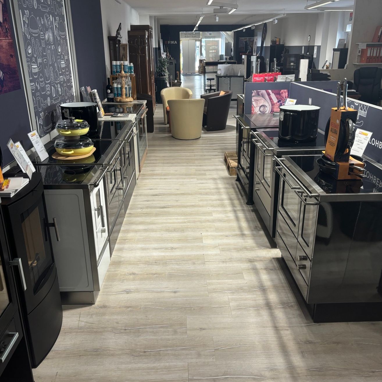 Eine moderne Küche mit schwarzen Geräten, hellen Holzböden und einer Kreidetafelwand. Stühle und Tische sind in der Nähe des Eingangs angeordnet, einige Artikel sind ausgestellt.