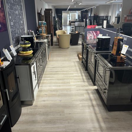 Eine moderne Küche mit schwarzen Geräten, hellen Holzböden und einer Kreidetafelwand. Stühle und Tische sind in der Nähe des Eingangs angeordnet, einige Artikel sind ausgestellt.