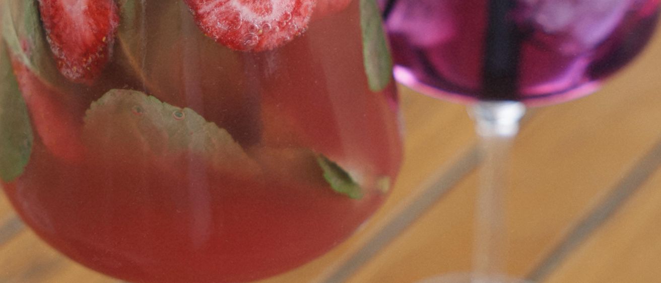 Zwei Weingläser mit farbenfrohen Cocktails, eines mit Erdbeeren und Minze, das andere mit Brombeeren, auf einem Holztisch.