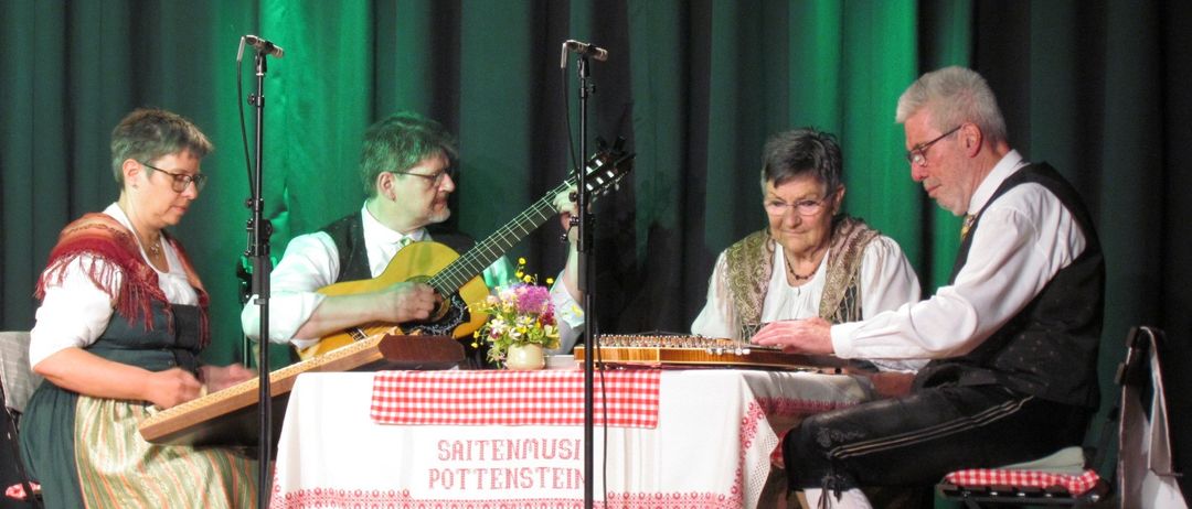 Vier Musiker spielen auf der Bühne. Einer spielt Gitarre, die anderen spielen Zithern. Ein Tisch mit einer rot-weiß karierten Tischdecke hat Blumen darauf. Der Hintergrund ist grün.