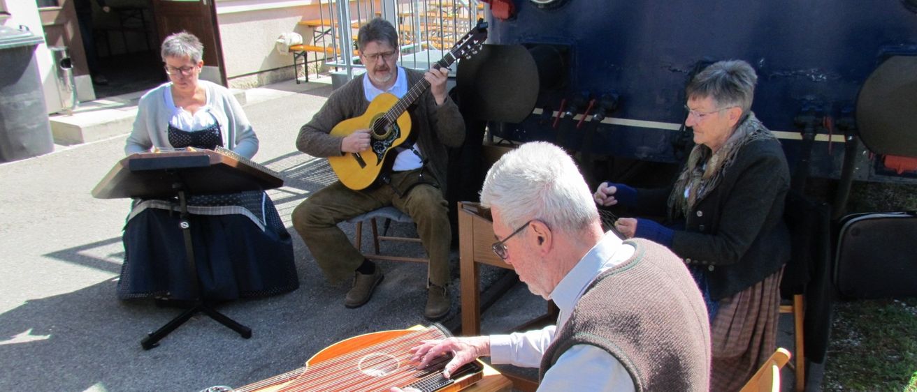 Vier ältere Erwachsene spielen Musik vor einem blauen Zug. Ein Mann spielt Gitarre, ein anderer Zither, eine Frau liest Noten und eine weitere Frau sitzt neben ihnen.
