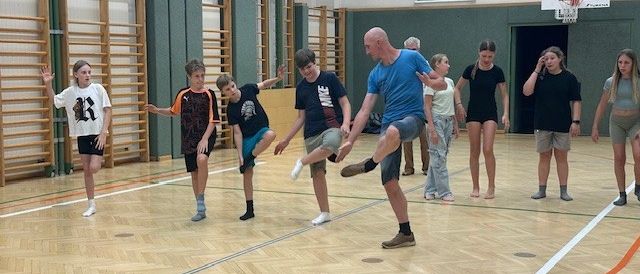 In einer Turnhalle nehmen eine Gruppe junger Menschen und Erwachsene an einem Tanz- oder Fitnesskurs teil. Ein Mann in Blau dehnt sich, während andere zusehen.
