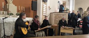 In einer Kirche spielen Musiker mit Gitarren und Zithern. Eine Statue und Kerzenständer schmücken den Hintergrund.