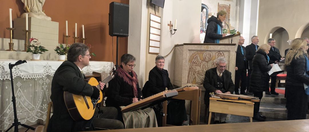 In einer Kirche spielen Musiker mit Gitarren und Zithern. Eine Statue und Kerzenständer schmücken den Hintergrund.