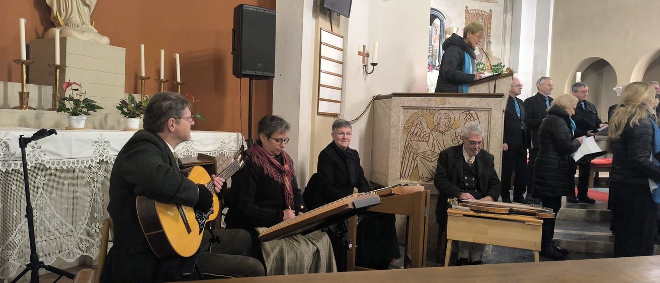 In einer Kirche spielen Musiker mit Gitarren und Zithern. Eine Statue und Kerzenständer schmücken den Hintergrund.