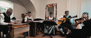 Vier Musiker in traditioneller Kleidung spielen Instrumente in einem Raum mit gewölbten Decken und gefliesten Böden. Zwei spielen Zither, und zwei spielen Gitarre.