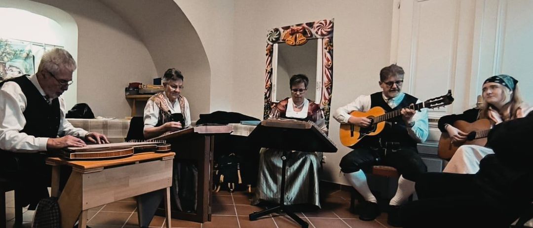 Vier Musiker in traditioneller Kleidung spielen Instrumente in einem Raum mit gewölbten Decken und gefliesten Böden. Zwei spielen Zither, und zwei spielen Gitarre.