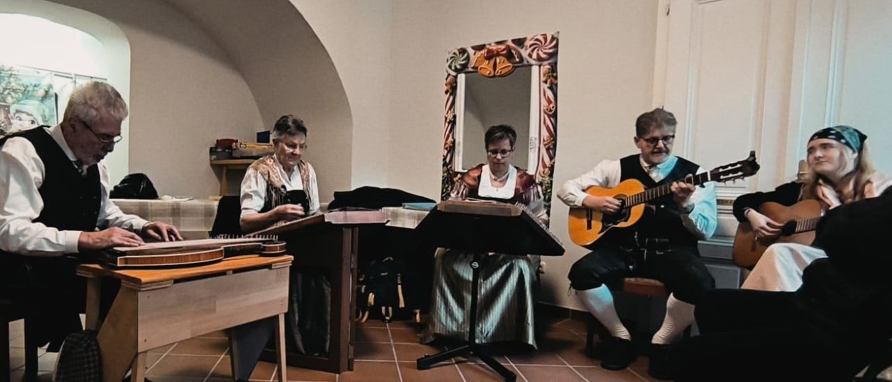 Vier Musiker in traditioneller Kleidung spielen Instrumente in einem Raum mit gewölbten Decken und gefliesten Böden. Zwei spielen Zither, und zwei spielen Gitarre.