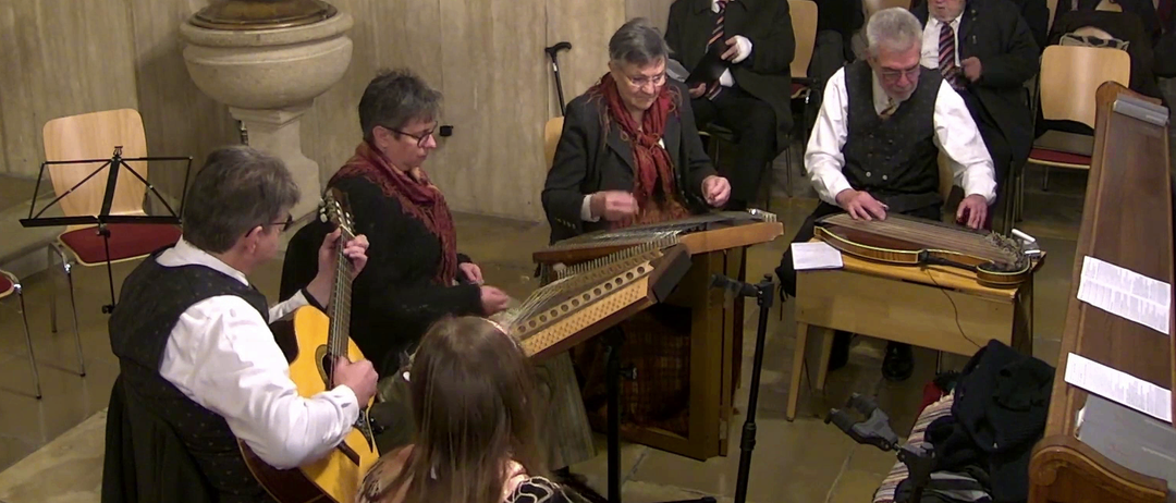 Eine Gruppe von Musikern in formeller Kleidung spielt Instrumente in einem Raum. Die Instrumente umfassen eine Gitarre und ein Saiteninstrument.
