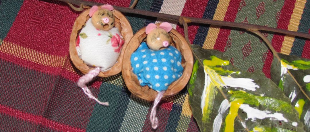 Zwei handgefertigte Mäuse, eine in Weiß und eine in Blau, befinden sich in Walnussschalen. Sie liegen auf einem Zweig mit Blättern auf einem bunten Tuch.