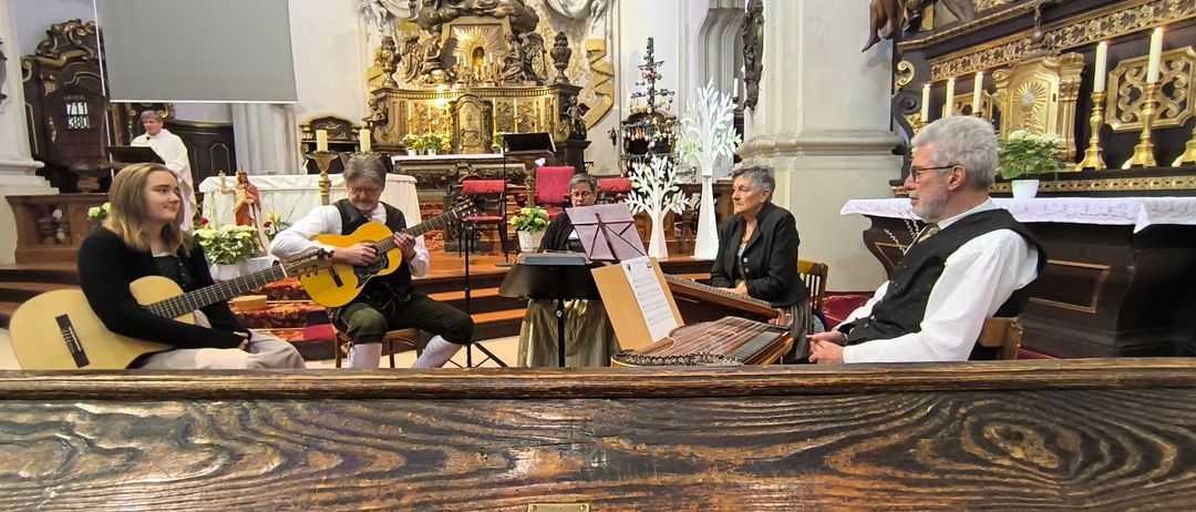 Eine Gruppe Musiker spielt Instrumente in einer Kirche. Ein Mann spielt Gitarre, eine Frau spielt Zither und eine andere liest Noten.
