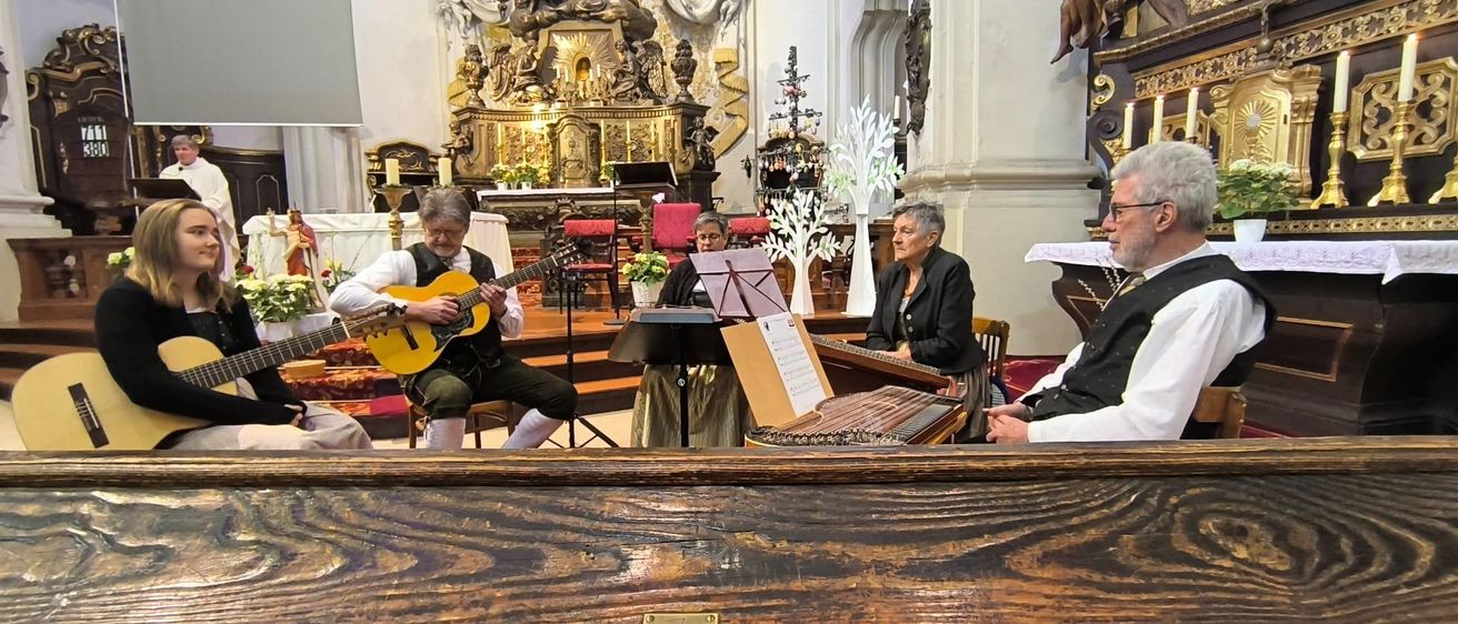 Eine Gruppe Musiker spielt Instrumente in einer Kirche. Ein Mann spielt Gitarre, eine Frau spielt Zither und eine andere liest Noten.