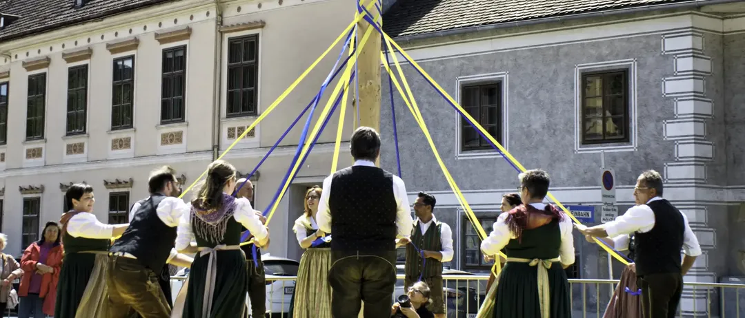 Mehrere Menschen versammeln sich um einen Maibaum, möglicherweise bei einem Fest. Ein Mann im Vest steht in der Mitte. Eine Frau hält eine Kamera und hält den Moment fest.