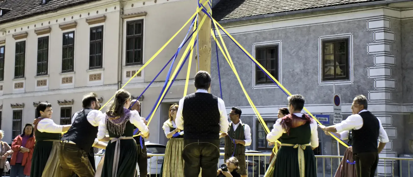 Mehrere Menschen versammeln sich um einen Maibaum, möglicherweise bei einem Fest. Ein Mann im Vest steht in der Mitte. Eine Frau hält eine Kamera und hält den Moment fest.