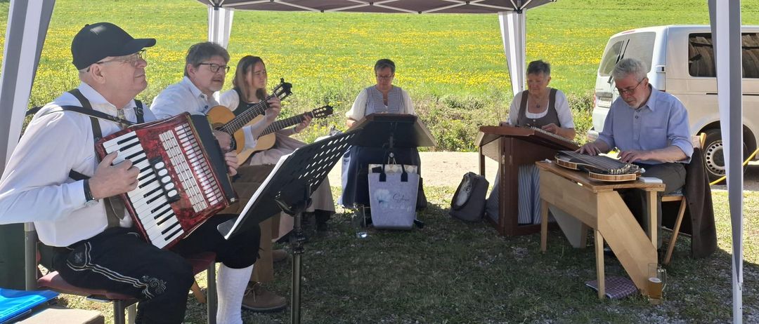 Eine Gruppe von Musikern spielt unter einem Zelt auf einer Wiese. Einige spielen Gitarren, andere Akkordeons und Harfen. Im Hintergrund sind ein weißer Van und Taschen zu sehen.